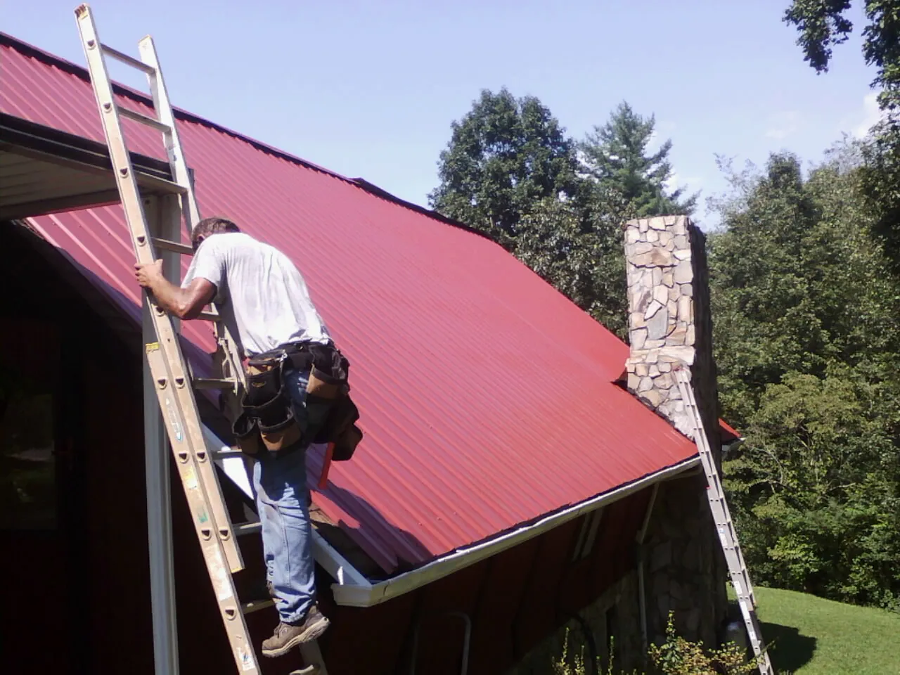 Roofer on a ladder beside a red metal roof and stone chimney — Soffit & Fascia Repair by Roofing Brothers in Catasauqua